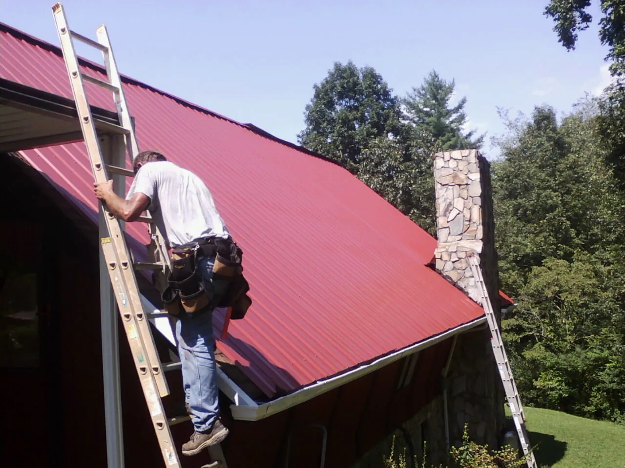 Roofer on a ladder beside a red metal roof and stone chimney — Soffit & Fascia Repair by Roofing Brothers in Summerfield
